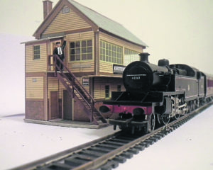 Black model train on tracks beside a model station with steps up the side and a model person stood at the top of the steps.